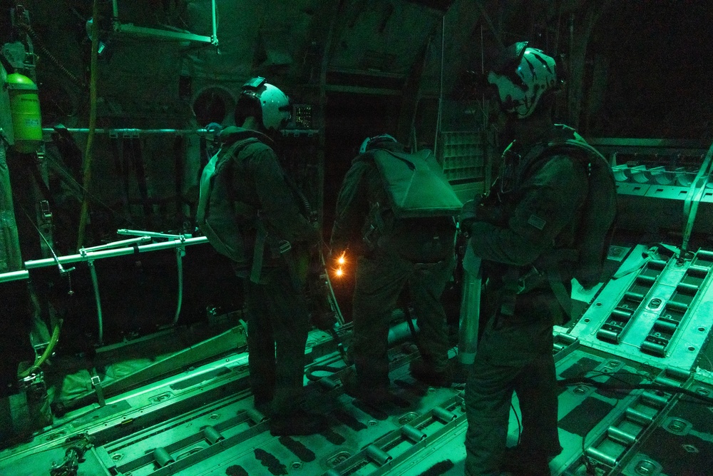 Three military personnel are standing inside what appears to be the cargo bay of a military transport aircraft, illuminated by green lighting. The individuals are equipped with parachute rigs and tactical helmets, suggesting preparation for a nighttime airborne operation. A small orange light source is visible between two of the personnel.