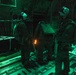 Three military personnel are standing inside what appears to be the cargo bay of a military transport aircraft, illuminated by green lighting. The individuals are equipped with parachute rigs and tactical helmets, suggesting preparation for a nighttime airborne operation. A small orange light source is visible between two of the personnel.