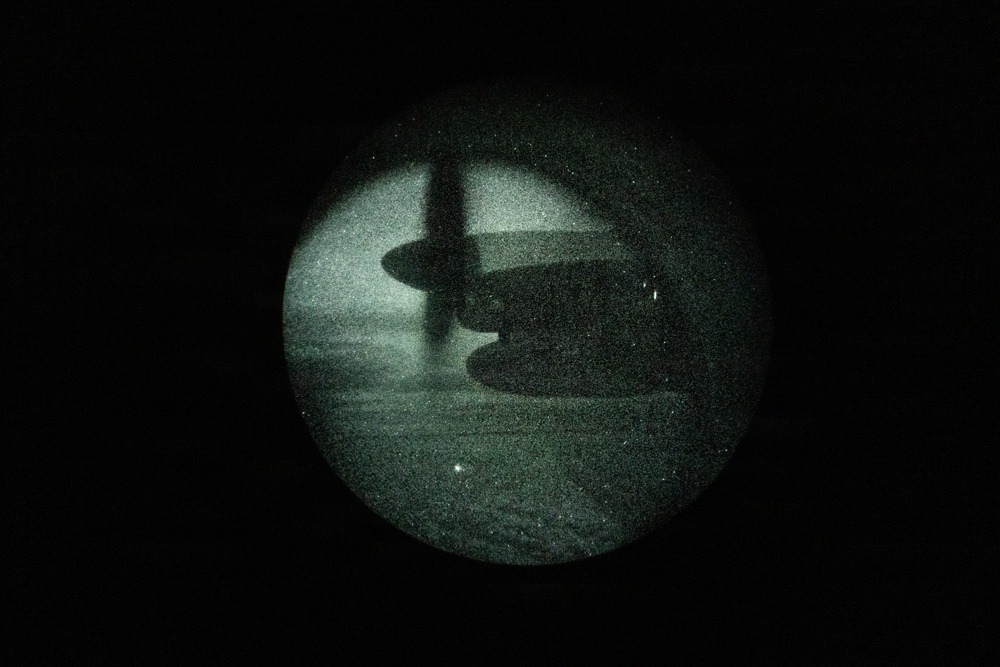 A night vision image captured through a circular optical device (likely night vision goggles or a scope) showing a large military transport aircraft on what appears to be a runway or tarmac. The green-tinted, grainy image is characteristic of Generation 2 or 3 image intensification night vision technology. The aircraft silhouette suggests a high-wing, multi-engine turboprop design consistent with a C-130 Hercules or similar military cargo aircraft.