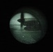 A night vision image captured through a circular optical device (likely night vision goggles or a scope) showing a large military transport aircraft on what appears to be a runway or tarmac. The green-tinted, grainy image is characteristic of Generation 2 or 3 image intensification night vision technology. The aircraft silhouette suggests a high-wing, multi-engine turboprop design consistent with a C-130 Hercules or similar military cargo aircraft.