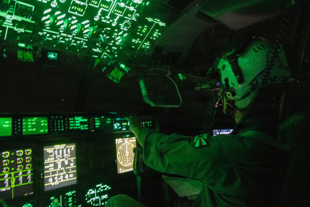 A military aircrew member is seated in a darkened cockpit illuminated entirely by green instrument lighting, consistent with night vision goggle (NVG) compatible lighting protocols. The cockpit features an extensive array of digital displays, overhead panels, and navigation screens typical of a large military transport aircraft. The green monochromatic lighting environment suggests a night operations mission.