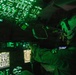 A military aircrew member is seated in a darkened cockpit illuminated entirely by green instrument lighting, consistent with night vision goggle (NVG) compatible lighting protocols. The cockpit features an extensive array of digital displays, overhead panels, and navigation screens typical of a large military transport aircraft. The green monochromatic lighting environment suggests a night operations mission.