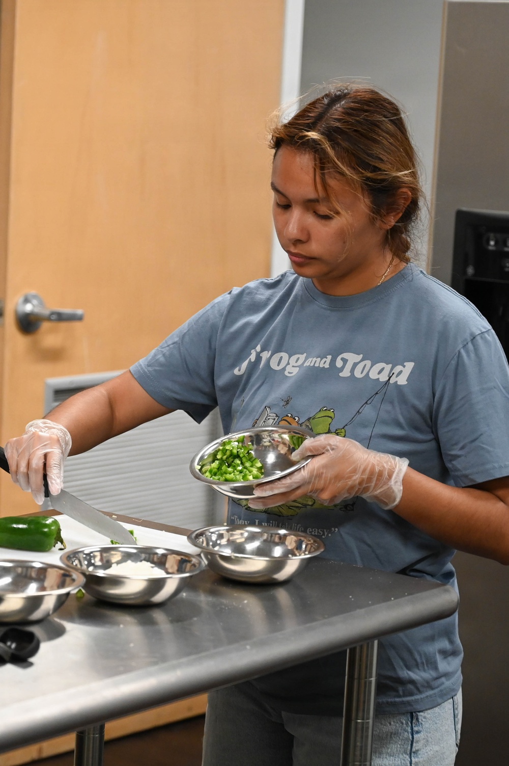 A young woman wearing a blue 'Frog and Toad' t-shirt and disposable gloves is preparing food in what appears to be a commercial or institutional kitchen. She is holding a stainless steel bowl containing chopped green bell peppers while using a knife on a metal prep table. Several additional stainless steel mixing bowls are arranged on the table in front of her.