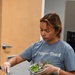 A young woman wearing a blue 'Frog and Toad' t-shirt and disposable gloves is preparing food in what appears to be a commercial or institutional kitchen. She is holding a stainless steel bowl containing chopped green bell peppers while using a knife on a metal prep table. Several additional stainless steel mixing bowls are arranged on the table in front of her.