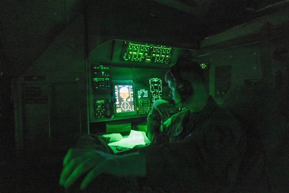 A military operator is seated at a darkened workstation illuminated primarily by green instrument lighting, consistent with night operations or low-light tactical environments. The station features multiple control panels, indicator lights, and at least one active display screen showing what appears to be a radar or sensor feed. The operator appears to be reviewing documents or a chart while monitoring the equipment.