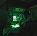 A military operator is seated at a darkened workstation illuminated primarily by green instrument lighting, consistent with night operations or low-light tactical environments. The station features multiple control panels, indicator lights, and at least one active display screen showing what appears to be a radar or sensor feed. The operator appears to be reviewing documents or a chart while monitoring the equipment.