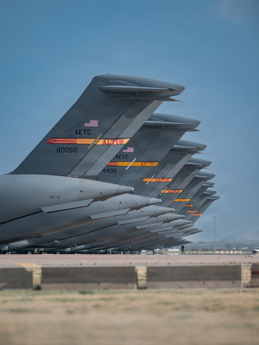 A row of U.S. Air Force military transport aircraft are lined up on a flight line, photographed from a low angle showing their vertical tail fins in perspective. The tails display AETC (Air Education and Training Command) markings with 'ALTUS' unit designations and American flag insignia, indicating these aircraft are assigned to Altus Air Force Base, Oklahoma. At least two serial numbers are legible — 80050 and 44135 — suggesting these are C-17 Globemaster III or similar large transport aircraft.