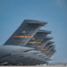A row of U.S. Air Force military transport aircraft are lined up on a flight line, photographed from a low angle showing their vertical tail fins in perspective. The tails display AETC (Air Education and Training Command) markings with 'ALTUS' unit designations and American flag insignia, indicating these aircraft are assigned to Altus Air Force Base, Oklahoma. At least two serial numbers are legible — 80050 and 44135 — suggesting these are C-17 Globemaster III or similar large transport aircraft.