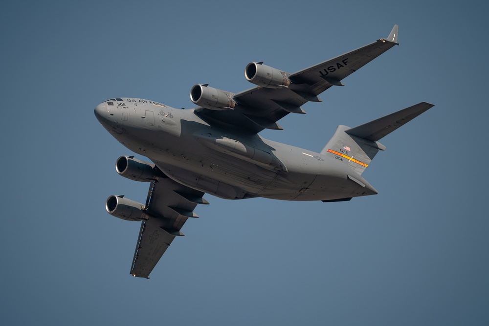 Two U.S. Air Force C-17 Globemaster III military transport aircraft are photographed in close formation flight against a clear blue sky. Both aircraft are painted in standard USAF gray livery and appear to be performing a demonstration or training flight. The aircraft are captured in a banking maneuver, revealing their undersides and wing configurations.