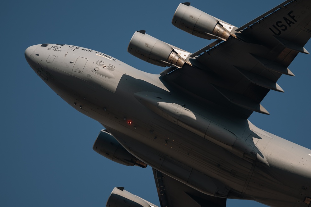 A close-up, low-angle photograph of a U.S. Air Force military transport aircraft in flight against a clear blue sky. The aircraft bears the markings '1102' and '97 AMW' (Air Mobility Wing), identifying it as a C-17 Globemaster III. The image captures the nose, fuselage, and multiple jet engines in sharp detail with dramatic perspective.