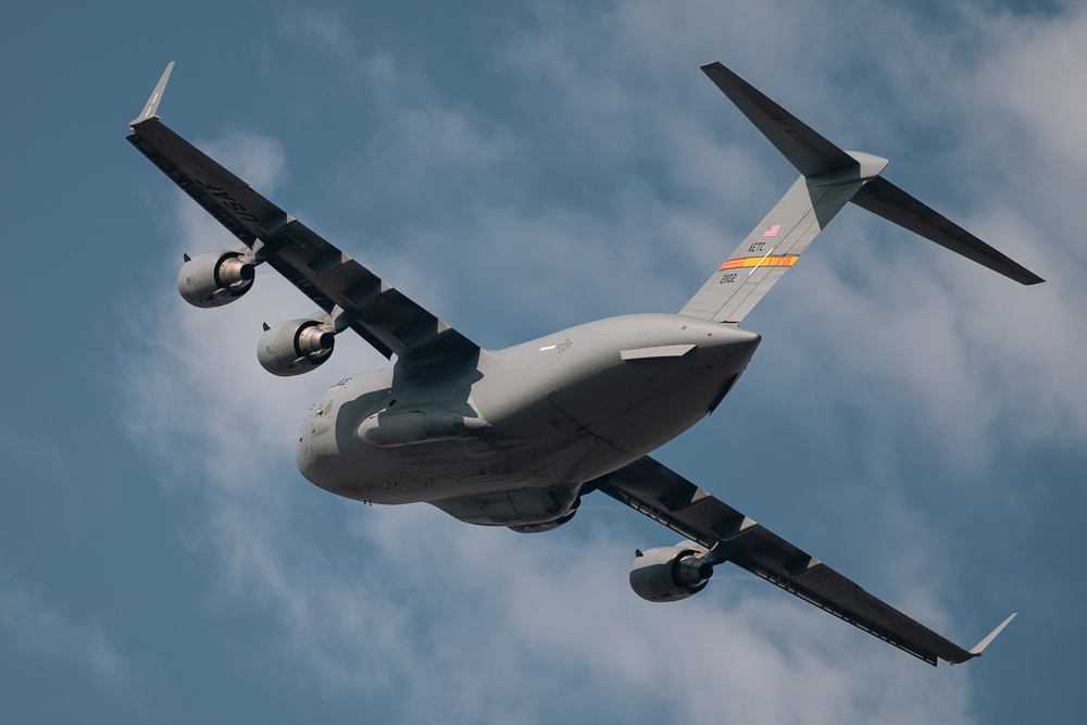 A large gray military transport aircraft, identifiable as a Boeing C-17 Globemaster III, is photographed from below at a steep banking angle against a partly cloudy blue sky. The aircraft displays USAF markings including an 'AETC' (Air Education and Training Command) tail band with orange and yellow colors, and the serial number '2102' is visible on the tail. The image appears to have been taken at an airshow or during a demonstration flight.