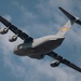 A large gray military transport aircraft, identifiable as a Boeing C-17 Globemaster III, is photographed from below at a steep banking angle against a partly cloudy blue sky. The aircraft displays USAF markings including an 'AETC' (Air Education and Training Command) tail band with orange and yellow colors, and the serial number '2102' is visible on the tail. The image appears to have been taken at an airshow or during a demonstration flight.