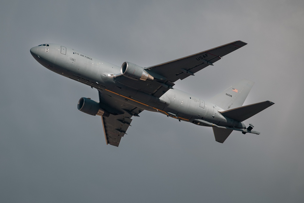 A U.S. Air Force military tanker aircraft, identified as a Boeing KC-46 Pegasus, is captured in flight against an overcast gray sky. The aircraft bears the serial number 66018 and displays 'U.S. AIR FORCE' markings along the fuselage. The aircraft appears to be climbing or in level flight with landing gear retracted.