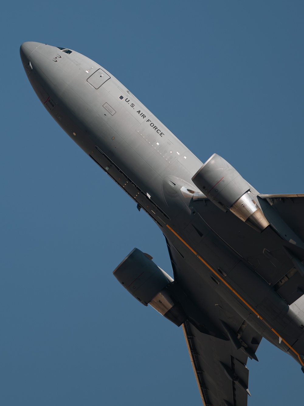 A U.S. Air Force military aircraft is photographed from a low angle during takeoff or low-altitude flight against a clear blue sky. The fuselage is painted in standard military gray with 'U.S. AIR FORCE' lettering clearly visible. The aircraft appears to be a large multi-engine jet, consistent with a tanker or transport platform such as a KC-46 Pegasus.