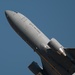 A U.S. Air Force military aircraft is photographed from a low angle during takeoff or low-altitude flight against a clear blue sky. The fuselage is painted in standard military gray with 'U.S. AIR FORCE' lettering clearly visible. The aircraft appears to be a large multi-engine jet, consistent with a tanker or transport platform such as a KC-46 Pegasus.