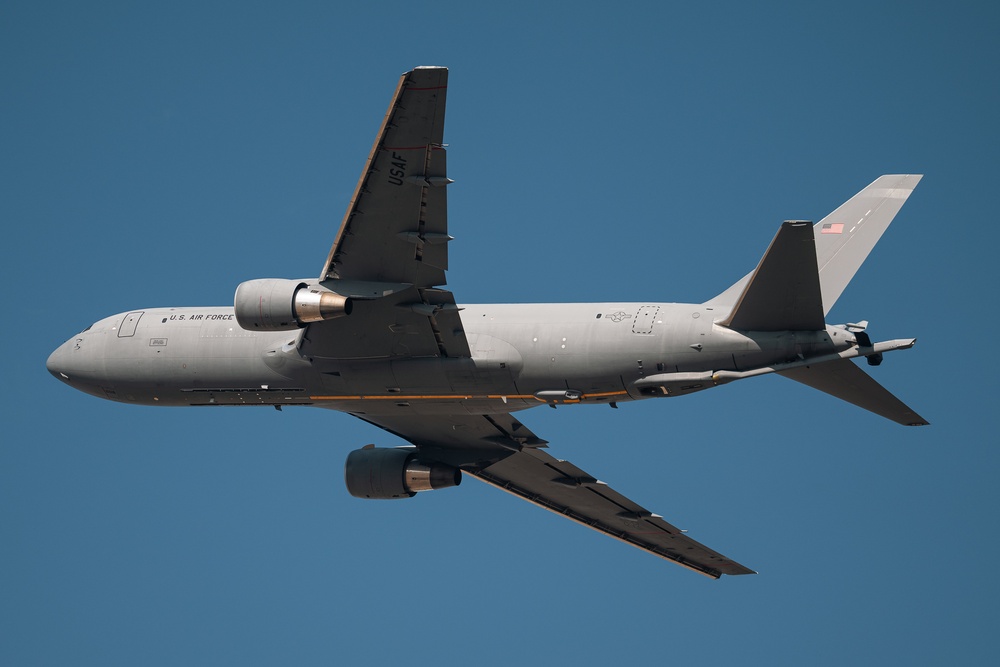 A U.S. Air Force military aircraft is photographed from below against a clear blue sky. The aircraft is painted in standard military gray livery and displays 'U.S. AIR FORCE' markings along the fuselage, along with a U.S. flag on the tail. Based on its twin-engine wide-body configuration and visible refueling boom housing, this appears to be a KC-46 Pegasus tanker aircraft.