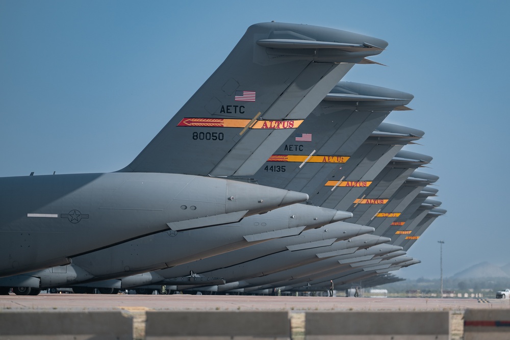 A row of U.S. Air Force military transport aircraft are lined up on a flight line, showing their tail sections in a repeating perspective. The aircraft are marked with 'AETC' (Air Education and Training Command) and 'ALTUS' insignia, indicating they are based at Altus Air Force Base, Oklahoma. Serial numbers visible include 80050 and 44135, consistent with C-17 Globemaster III aircraft.