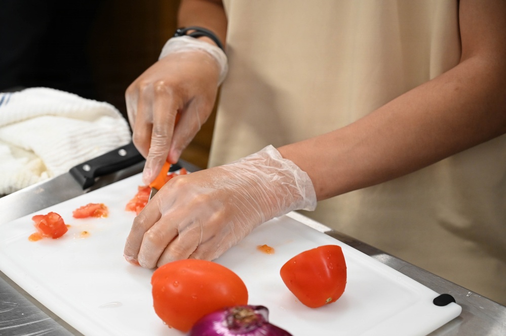 A person wearing disposable plastic gloves and a beige apron is chopping tomatoes on a white cutting board. The scene appears to be in a professional or semi-professional kitchen setting, with stainless steel surfaces visible. Two whole tomatoes and a partial red onion are also present on the cutting board.