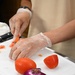 A person wearing disposable plastic gloves and a beige apron is chopping tomatoes on a white cutting board. The scene appears to be in a professional or semi-professional kitchen setting, with stainless steel surfaces visible. Two whole tomatoes and a partial red onion are also present on the cutting board.