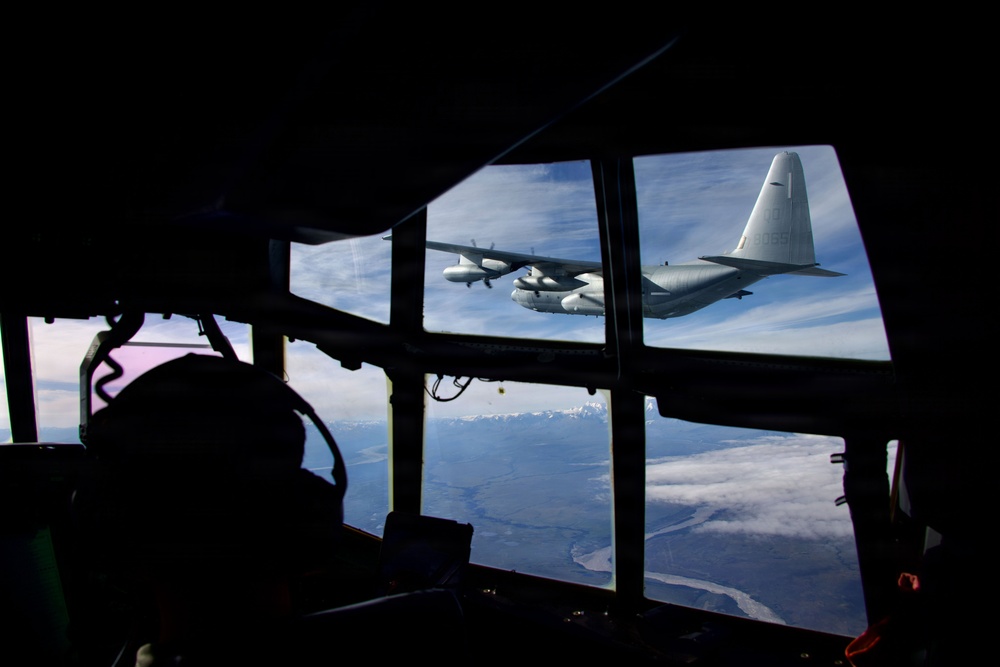 The image is taken from inside a military aircraft cockpit looking through the multi-pane windshield at another military turboprop aircraft flying in close formation. The exterior aircraft appears to be a Lockheed C-130 Hercules, identifiable by its distinctive high-wing, four-turboprop configuration and T-tail design. Below, a landscape with snow-capped mountains, a river valley, and partial cloud cover is visible.