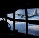 The image is taken from inside a military aircraft cockpit looking through the multi-pane windshield at another military turboprop aircraft flying in close formation. The exterior aircraft appears to be a Lockheed C-130 Hercules, identifiable by its distinctive high-wing, four-turboprop configuration and T-tail design. Below, a landscape with snow-capped mountains, a river valley, and partial cloud cover is visible.
