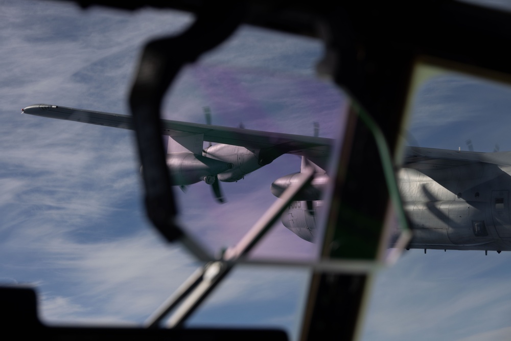 The image is taken from inside a military aircraft cockpit, looking through the windshield frame at another large military turboprop aircraft flying in close formation. The subject aircraft appears to be a C-130 Hercules or similar four-engine turboprop transport, photographed at altitude with a blue sky and cloud layer visible in the background. The cockpit framing and window reflections create a layered, partially obscured perspective.