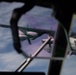 The image is taken from inside a military aircraft cockpit, looking through the windshield frame at another large military turboprop aircraft flying in close formation. The subject aircraft appears to be a C-130 Hercules or similar four-engine turboprop transport, photographed at altitude with a blue sky and cloud layer visible in the background. The cockpit framing and window reflections create a layered, partially obscured perspective.
