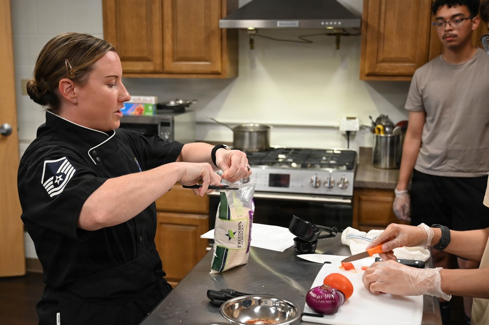 A military service member wearing a black chef's coat with a Master Sergeant rank insignia is conducting a cooking class or demonstration in a kitchen setting. She is opening a bag labeled 'Freedom's Choice' while another person in gloves chops carrots on a cutting board. A third individual, a young man in a gray t-shirt, observes in the background.