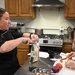 A military service member wearing a black chef's coat with a Master Sergeant rank insignia is conducting a cooking class or demonstration in a kitchen setting. She is opening a bag labeled 'Freedom's Choice' while another person in gloves chops carrots on a cutting board. A third individual, a young man in a gray t-shirt, observes in the background.