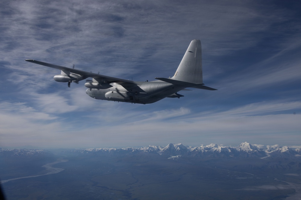 A military turboprop aircraft, consistent with a C-130 Hercules variant, is photographed in flight from another aircraft at altitude. The aircraft is painted in a gray/silver scheme and is flying over a vast landscape with snow-capped mountain ranges visible in the background. The sky is partly cloudy with high cirrus clouds.