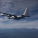 A military turboprop aircraft, consistent with a C-130 Hercules variant, is photographed in flight from another aircraft at altitude. The aircraft is painted in a gray/silver scheme and is flying over a vast landscape with snow-capped mountain ranges visible in the background. The sky is partly cloudy with high cirrus clouds.