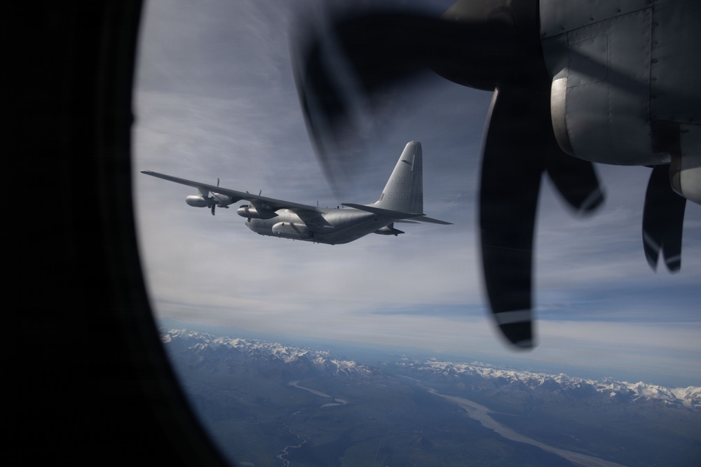 The image is taken from inside a military turboprop aircraft through a circular porthole window, showing a second similar aircraft flying in formation. A propeller from the observer aircraft is visible in the foreground on the right side. Snow-capped mountain ranges and a river valley are visible far below, suggesting high-altitude flight.