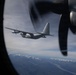 The image is taken from inside a military turboprop aircraft through a circular porthole window, showing a second similar aircraft flying in formation. A propeller from the observer aircraft is visible in the foreground on the right side. Snow-capped mountain ranges and a river valley are visible far below, suggesting high-altitude flight.