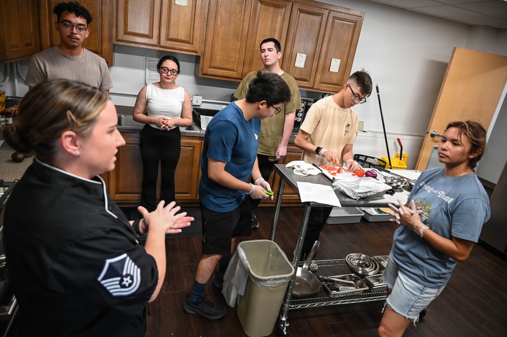 A military instructor wearing a black chef uniform with a Master Sergeant rank insignia is conducting a cooking class or culinary training session with a group of young individuals. Participants are gathered around a stainless steel prep cart where tomatoes and other ingredients are being chopped. The instructor appears to be giving verbal instructions while demonstrating or explaining food preparation techniques.