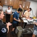 A military instructor wearing a black chef uniform with a Master Sergeant rank insignia is conducting a cooking class or culinary training session with a group of young individuals. Participants are gathered around a stainless steel prep cart where tomatoes and other ingredients are being chopped. The instructor appears to be giving verbal instructions while demonstrating or explaining food preparation techniques.