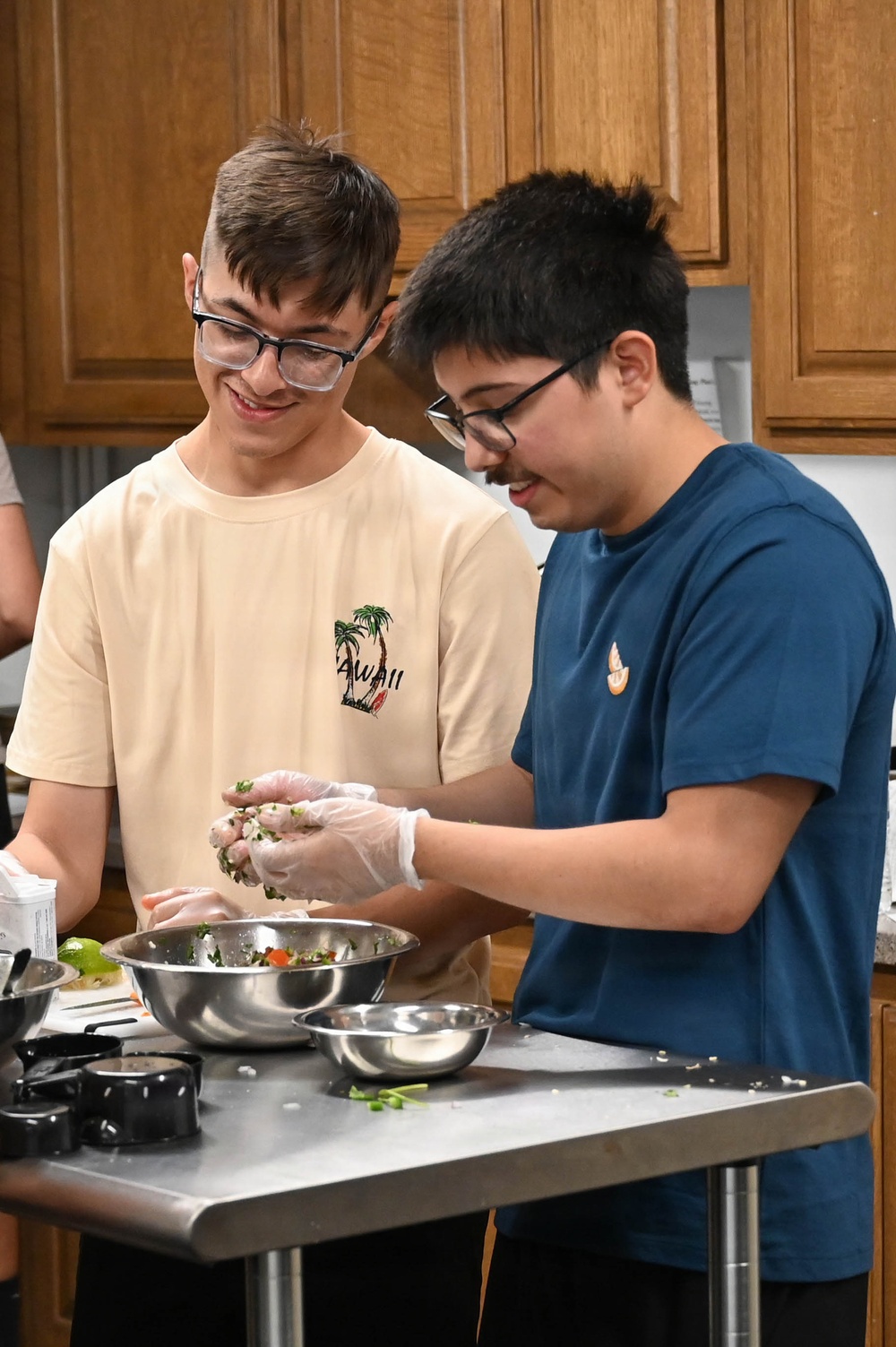 Two teenage boys wearing glasses are working together at a stainless steel prep table in what appears to be a kitchen classroom or culinary education setting. Both are smiling and engaged in mixing or handling food ingredients in metal mixing bowls. The boy on the right is wearing disposable plastic gloves and appears to be mixing a dish containing herbs and vegetables.