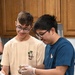 Two teenage boys wearing glasses are working together at a stainless steel prep table in what appears to be a kitchen classroom or culinary education setting. Both are smiling and engaged in mixing or handling food ingredients in metal mixing bowls. The boy on the right is wearing disposable plastic gloves and appears to be mixing a dish containing herbs and vegetables.