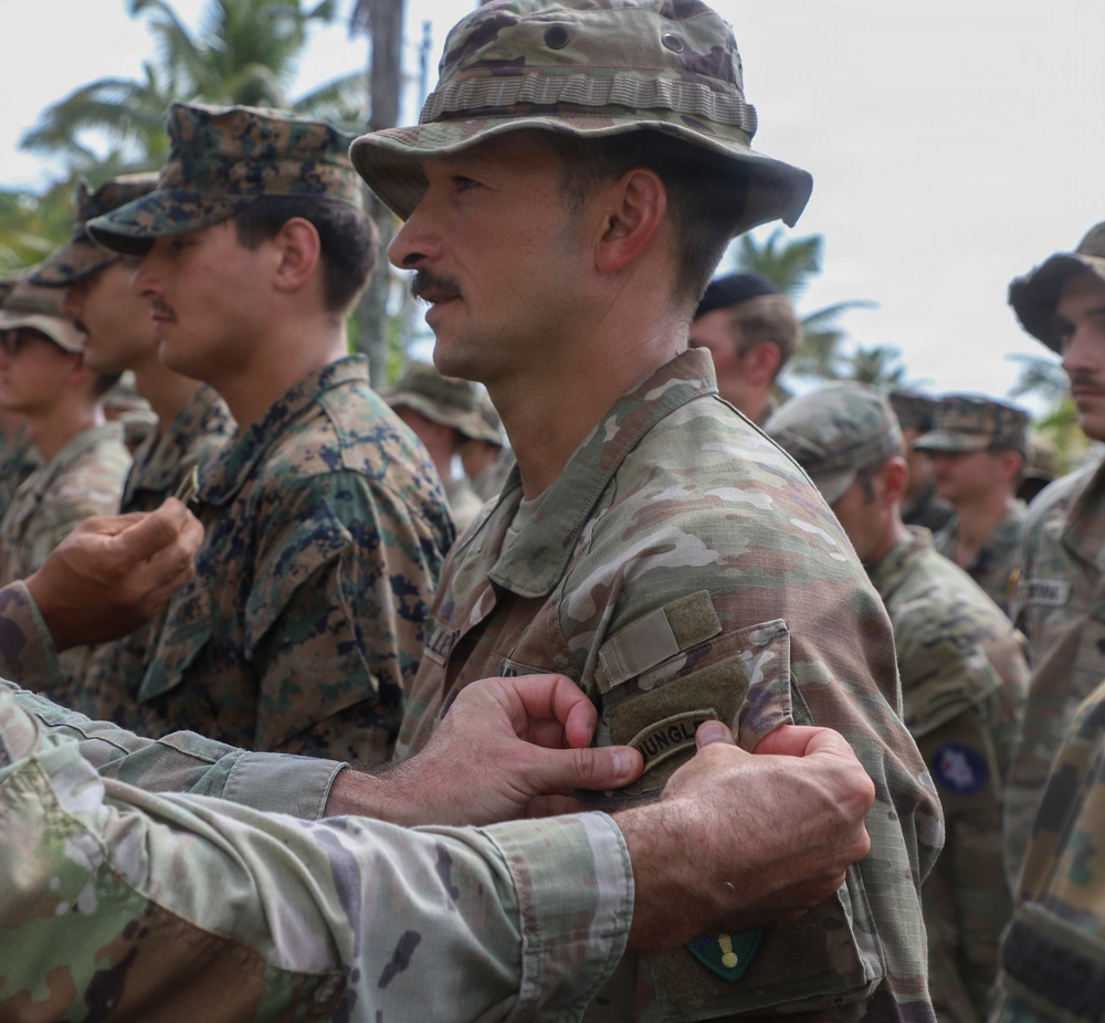 A military ceremony appears to be taking place outdoors in a tropical setting, where a patch is being affixed to a soldier's uniform sleeve. The central figure wears MultiCam/OCP camouflage and a boonie hat, with a visible 'JUNGLE' tab being attached to his left shoulder. Multiple service members in mixed uniforms, including USMC MARPAT and Army OCP, are present in formation.