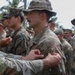 A military ceremony appears to be taking place outdoors in a tropical setting, where a patch is being affixed to a soldier's uniform sleeve. The central figure wears MultiCam/OCP camouflage and a boonie hat, with a visible 'JUNGLE' tab being attached to his left shoulder. Multiple service members in mixed uniforms, including USMC MARPAT and Army OCP, are present in formation.