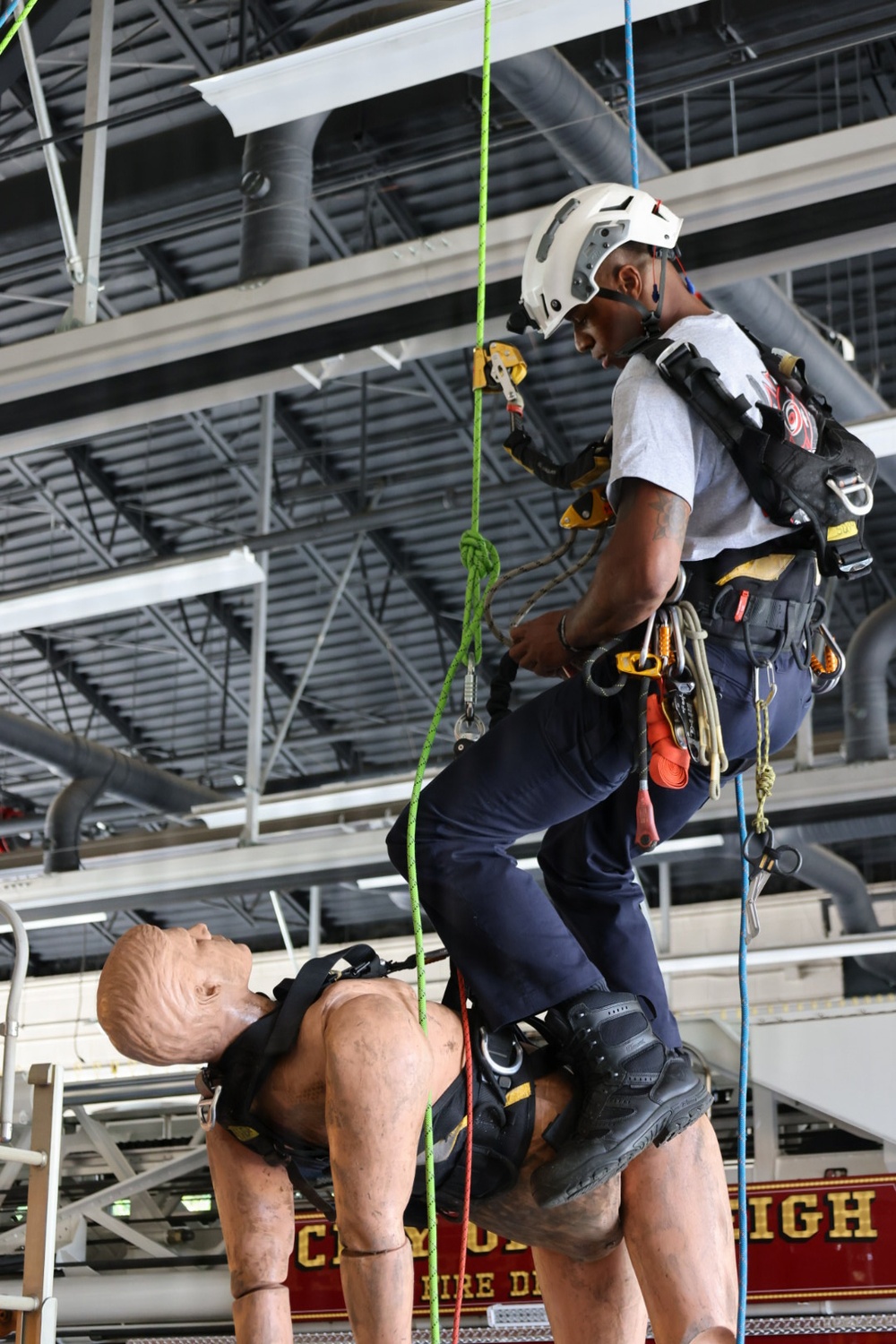 A firefighter or rescue technician in full rope rescue harness and white helmet is suspended on ropes inside what appears to be a fire station training facility. The person is performing a rope rescue drill, managing a training dummy (rescue mannequin) that is also suspended by ropes and harness. Partial text on a fire apparatus in the background reads 'FIRE D[EPARTMENT]' and 'IGH', suggesting a city fire department facility.