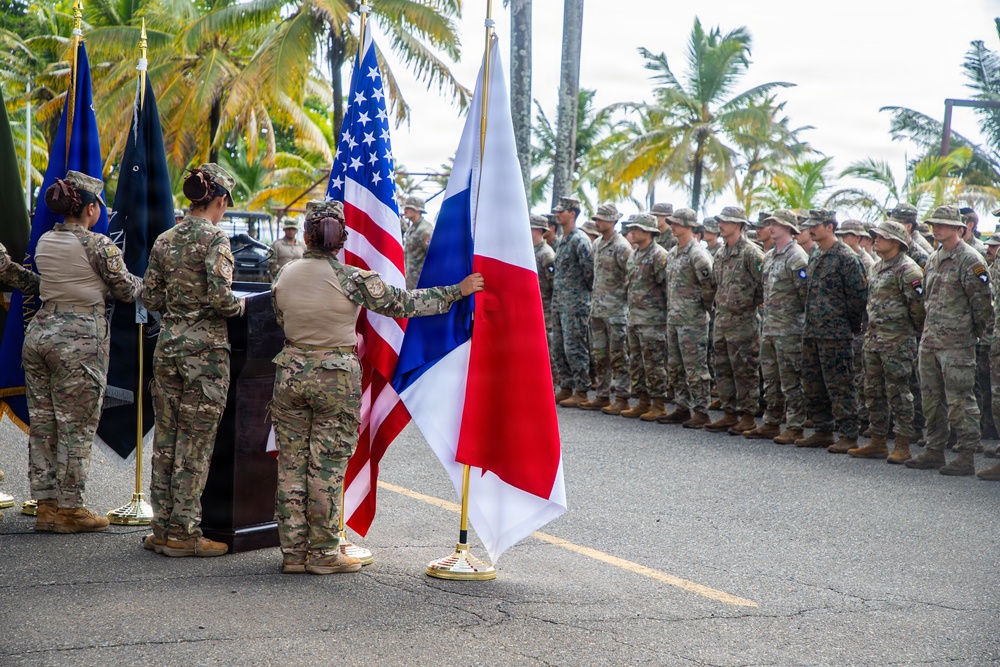 A military ceremony is taking place outdoors in a tropical setting, featuring soldiers in camouflage uniforms arranged in formation. Two national flags are prominently displayed: the United States flag and what appears to be the flag of Panama, suggesting a joint military event or bilateral ceremony. Several female soldiers are handling the flags near a podium while a large formation of troops stands at attention in the background.