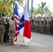 A military ceremony is taking place outdoors in a tropical setting, featuring soldiers in camouflage uniforms arranged in formation. Two national flags are prominently displayed: the United States flag and what appears to be the flag of Panama, suggesting a joint military event or bilateral ceremony. Several female soldiers are handling the flags near a podium while a large formation of troops stands at attention in the background.