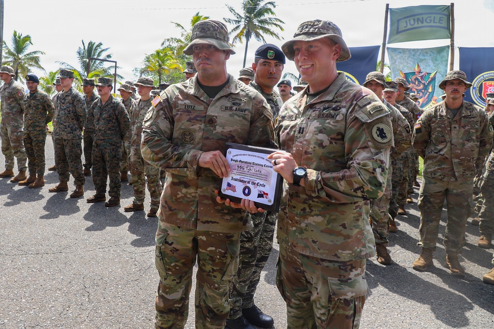 Two U.S. Army soldiers are holding a certificate from the Jungle Operations Training Course, with one identified as SSG Tyler Lutz receiving an 'Instructor of the Cycle' award. The ceremony is taking place outdoors in a tropical setting with a large formation of multinational military personnel standing at attention in the background. A green 'JUNGLE' banner and military insignia flags are visible in the background.