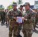 Two U.S. Army soldiers are holding a certificate from the Jungle Operations Training Course, with one identified as SSG Tyler Lutz receiving an 'Instructor of the Cycle' award. The ceremony is taking place outdoors in a tropical setting with a large formation of multinational military personnel standing at attention in the background. A green 'JUNGLE' banner and military insignia flags are visible in the background.