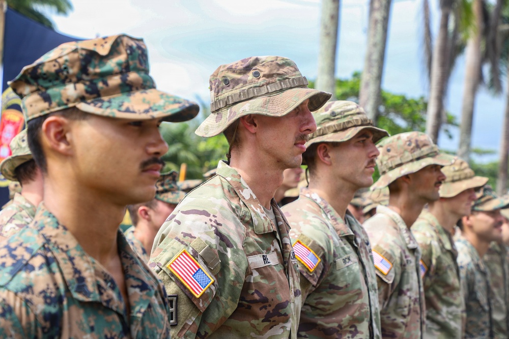 A formation of military personnel standing at attention in what appears to be an outdoor ceremony or parade. The soldiers are wearing different camouflage uniforms, with most displaying U.S. flag patches on their sleeves. The setting appears to be a tropical location, evidenced by palm trees visible in the background.