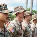 A formation of military personnel standing at attention in what appears to be an outdoor ceremony or parade. The soldiers are wearing different camouflage uniforms, with most displaying U.S. flag patches on their sleeves. The setting appears to be a tropical location, evidenced by palm trees visible in the background.