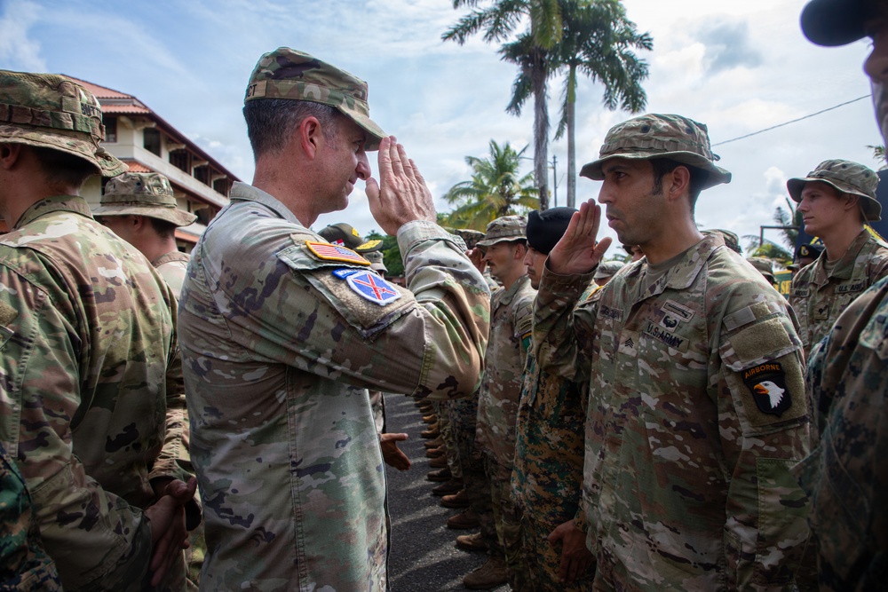 A U.S. Army officer and a sergeant exchange salutes during what appears to be a formal military ceremony or inspection. Multiple soldiers in OCP (Operational Camouflage Pattern) uniforms are standing in formation. The setting appears to be a tropical location based on the palm trees and vegetation visible in the background.