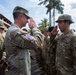 A U.S. Army officer and a sergeant exchange salutes during what appears to be a formal military ceremony or inspection. Multiple soldiers in OCP (Operational Camouflage Pattern) uniforms are standing in formation. The setting appears to be a tropical location based on the palm trees and vegetation visible in the background.