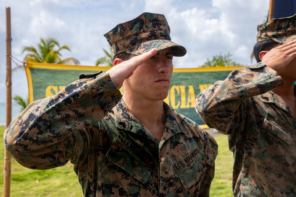 Two U.S. Marines in MARPAT digital camouflage uniforms are rendering a salute outdoors. The foreground Marine's 'U.S. MARINES' chest tape is clearly visible. The setting appears to be a tropical location, suggested by palm trees and a green-and-yellow banner in the background.