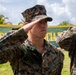Two U.S. Marines in MARPAT digital camouflage uniforms are rendering a salute outdoors. The foreground Marine's 'U.S. MARINES' chest tape is clearly visible. The setting appears to be a tropical location, suggested by palm trees and a green-and-yellow banner in the background.